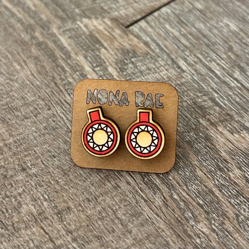 May include: A pair of wooden earrings with a red and white sun design. The earrings are on a card that says "Nona Rae".