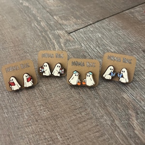 May include: Four pairs of wooden ghost earrings on brown cards. Each pair features a white ghost with black eyes and a unique accessory: a book, books, a trick-or-treat bucket, or a hat. The cards are labeled "NONA RAE".