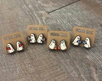 May include: Four pairs of wooden ghost earrings on brown cards. Each pair features a white ghost with black eyes and a unique accessory: a book, books, a trick-or-treat bucket, or a hat. The cards are labeled "NONA RAE".