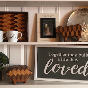 May include: A white shelf with two shelves. The top shelf has a wooden tray with a geometric pattern, two white mugs with black lettering that says "wifey" and "hubby", a framed photo with a white background and black text that says "of all the walks we've taken this was my favorite", a wooden cow figurine with a geometric pattern, and a silver metal tray. The bottom shelf has a small potted succulent plant, a wooden piggy bank with a geometric pattern, and a framed sign with a gray background and white text that says "Together they built a life they loved".