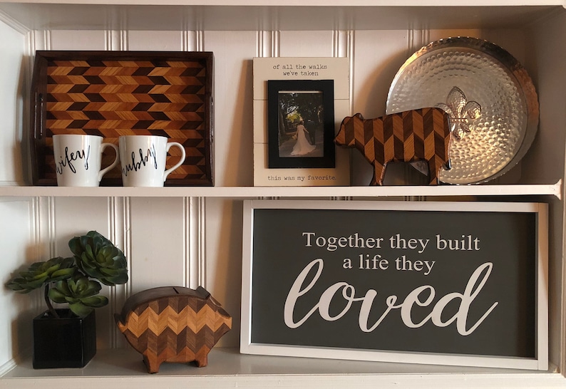 May include: A white shelf with two shelves. The top shelf has a wooden tray with a geometric pattern, two white mugs with black lettering that says "wifey" and "hubby", a framed photo with a white background and black text that says "of all the walks we've taken this was my favorite", a wooden cow figurine with a geometric pattern, and a silver metal tray. The bottom shelf has a small potted succulent plant, a wooden piggy bank with a geometric pattern, and a framed sign with a gray background and white text that says "Together they built a life they loved".