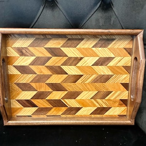 May include: A rectangular wooden serving tray with a geometric pattern. The tray features a dark brown frame and handles, with a central design of alternating light and dark wood chevrons. The tray is designed for serving food or drinks.