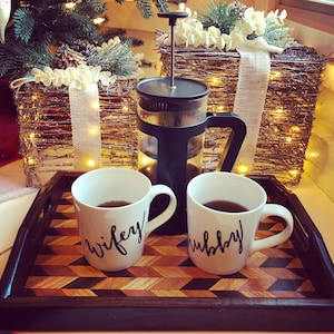 May include: Two white ceramic mugs with black lettering on a wooden tray. The mugs say "wifey" and "hubby". A french press coffee maker is in the background.