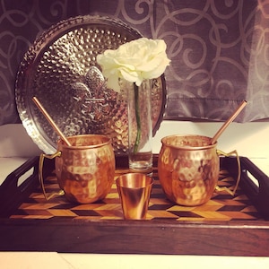 May include: Two copper mugs with brass handles and a small copper shot glass on a wooden tray with a patterned inlay. A single white flower sits in a clear glass vase on the tray.