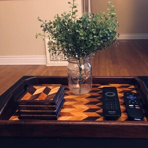 May include: A wooden tray with a geometric pattern, featuring a stack of four wooden coasters and two remote controls. A clear glass jar with a green plant is in the background.