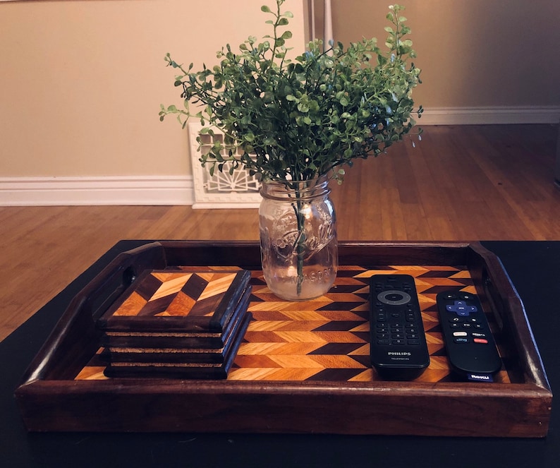 May include: A wooden tray with a geometric pattern, featuring a stack of four wooden coasters and two remote controls. A clear glass jar with a green plant is in the background.