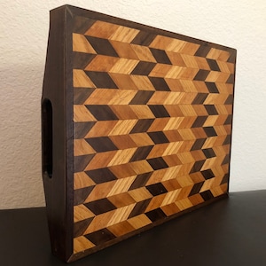 May include: A wooden cutting board with a geometric pattern made of different shades of wood. The board has a handle on the side and is made of walnut and maple wood.