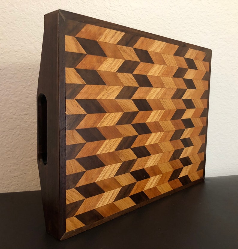 May include: A wooden cutting board with a geometric pattern made of different shades of wood. The board has a handle on the side and is made of walnut and maple wood.