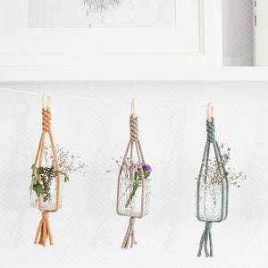 Könnte beinhalten: Drei Makramee-Pflanzenhänger mit Glasbehältern und kleinen Blumensträußen. Die Hänger sind aus naturfarbenem Kordel gefertigt und haben oben Holzringe. Die Behälter sind aus klarem Glas und haben eine strukturierte Oberfläche.