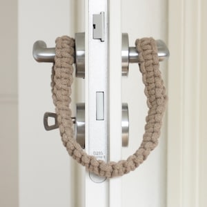 Könnte beinhalten: Ein beigefarbener gestrickter Türgriffbezug ist um einen Türgriff gewickelt. Der Griff ist silberfarben und hat ein Schlüsselloch. Die Tür ist weiß.