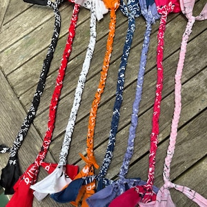 Puede incluir: Una colección de juguetes trenzados para perros hechos de pañuelos en varios colores, incluyendo negro, rojo, blanco, naranja, azul marino, morado y rosa. Cada juguete tiene un asa trenzada y un pañuelo al final.