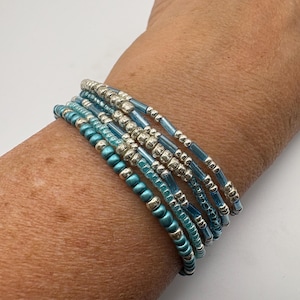 May include: A stack of beaded bracelets in shades of teal and silver. The bracelets feature a mix of small, round beads and larger, metallic silver beads. The bracelets are worn on a wrist, showcasing the jewelry's design and color.