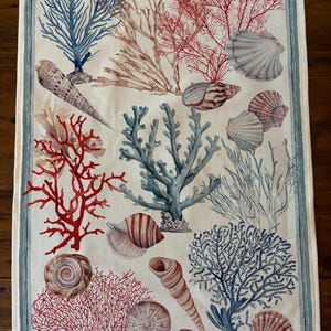 May include: A rectangular white tea towel with a blue border. The towel features watercolour illustrations of seashells, coral, and seaweed in shades of red, blue, and brown. The design is a coastal theme.