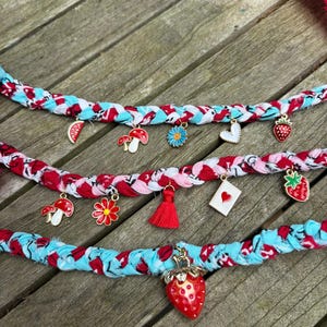May include: Three braided fabric necklaces with red, white, and blue patterns. Each necklace is adorned with various charms, including strawberries, mushrooms, flowers, and hearts. The necklaces are displayed on a wooden surface.