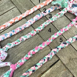 May include: Five braided fabric dog toys in various colors. The toys are made of colorful patterned fabric, including floral and geometric designs. Each toy has a different color scheme, with pink, blue, orange, and green accents. The toys are arranged diagonally on a wooden surface.