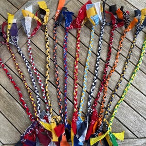 May include: A collection of ten braided dog toys made from colorful patterned fabric strips. Each toy features a long, tightly woven braid with loose, frayed fabric ends in various colors including red, blue, yellow, orange, black, and green.