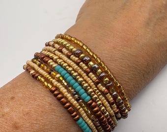 May include: A stack of beaded bracelets in shades of gold, brown, and turquoise. The bracelets are made of small, round beads and are stacked on top of each other. The bracelets are on a wrist.