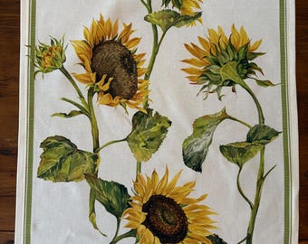 Op de afbeelding: Een witte rechthoekige theedoek met een botanische illustratie van vier zonnebloemen met gele bloemblaadjes, donkerbruine harten en groene bladeren. De doek is omlijst door een dunne groene rand met verticale strepen langs de randen.