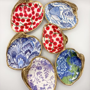 May include: Six decorative oyster shell dishes with gold-painted edges. The interiors feature various patterns, including red tomatoes on white, blue floral designs, and a blue and white seashell motif. These unique dishes add a touch of coastal charm.