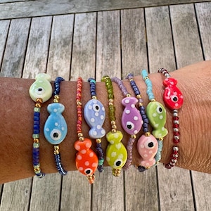 Op de afbeelding: Een collectie van acht kralenarmbanden met keramische visvormige bedels. Elke armband is gemaakt met kleine, kleurrijke glaskralen en goudkleurige metalen tussenstukjes. De visbedels zijn beschilderd in diverse pastel- en felle kleuren.