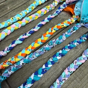 Könnte beinhalten: Nahaufnahme von mehreren geflochtenen Stoff-Hundespielzeugen in verschiedenen Farben. Die Spielzeuge bestehen aus Stoffstreifen mit Mustern, darunter Blau, Gelb, Rosa und Orange. Die Zöpfe sind diagonal auf einer Holzoberfläche angeordnet.