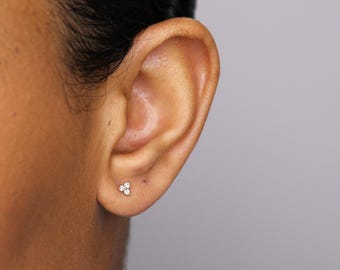 Puede incluir: Primer plano de la oreja de una persona con un pequeño pendiente de diamante de tres piedras. El pendiente está engastado en un delicado engaste de oro.