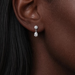 Puede incluir: Primer plano de una oreja adornada con pendientes colgantes de diamantes. Cada pendiente presenta un diamante redondo y un colgante de diamante en forma de pera. Los pendientes est&aacute;n engarzados en un metal plateado.