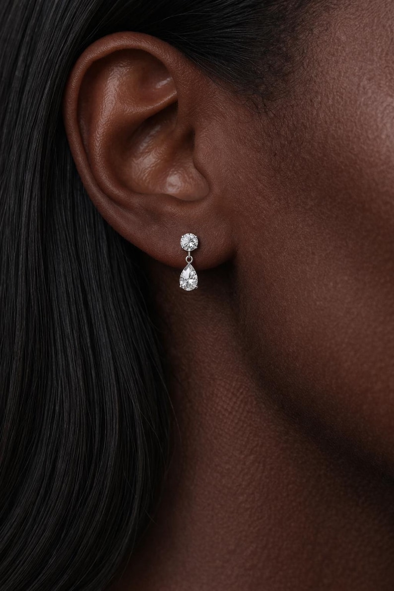 Puede incluir: Primer plano de una oreja adornada con pendientes colgantes de diamantes. Cada pendiente presenta un diamante redondo y un colgante de diamante en forma de pera. Los pendientes est&aacute;n engarzados en un metal plateado.