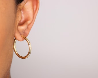 May include: A close-up of a gold hoop earring on an ear. The earring is a simple, circular design with a polished finish. The earring is made of gold-coloured metal and reflects light. The background is a soft, neutral colour.