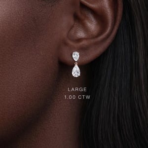 May include: A close-up of a person's ear with a pair of diamond drop earrings. The earrings have a pear-shaped design, with a smaller diamond above a larger one. The text "LARGE 1.00 CTW" is visible.