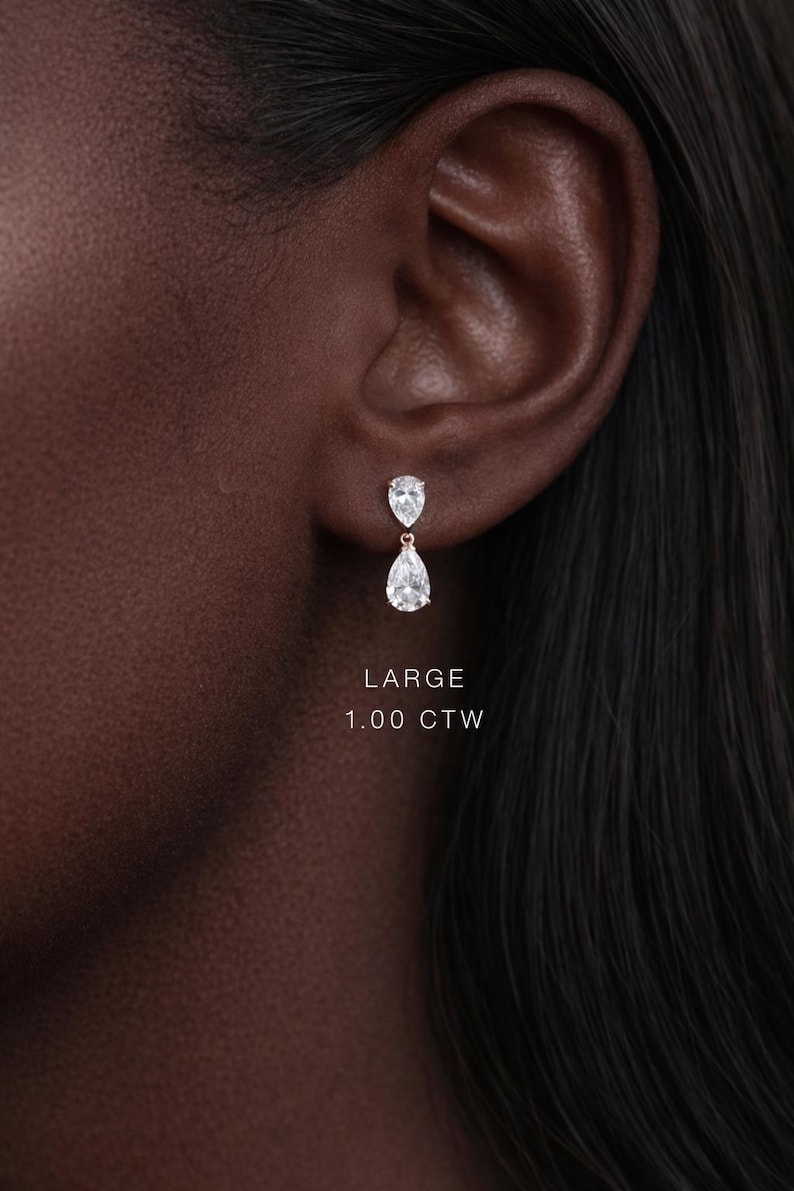 May include: A close-up of a person's ear with a pair of diamond drop earrings. The earrings have a pear-shaped design, with a smaller diamond above a larger one. The text "LARGE 1.00 CTW" is visible.