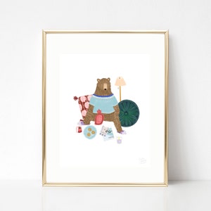 May include: A brown bear wearing a blue sweater sits on a pink and red patterned pillow with a green and gold round cushion behind it. The bear is holding a red hot water bottle and has a cup of tea, a plate of cookies, and a pair of glasses in front of it. There is a lamp with a yellow shade in the background.
