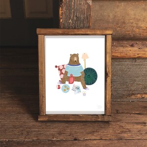 May include: A brown bear wearing a blue sweater and a beaded necklace sits on a patterned pillow with a teal blue round cushion behind it. The bear is holding a red hot water bottle and a glass of water. There are cookies, a red and white striped cup, a blue and white patterned map, and a lamp with a white shade on the white background.
