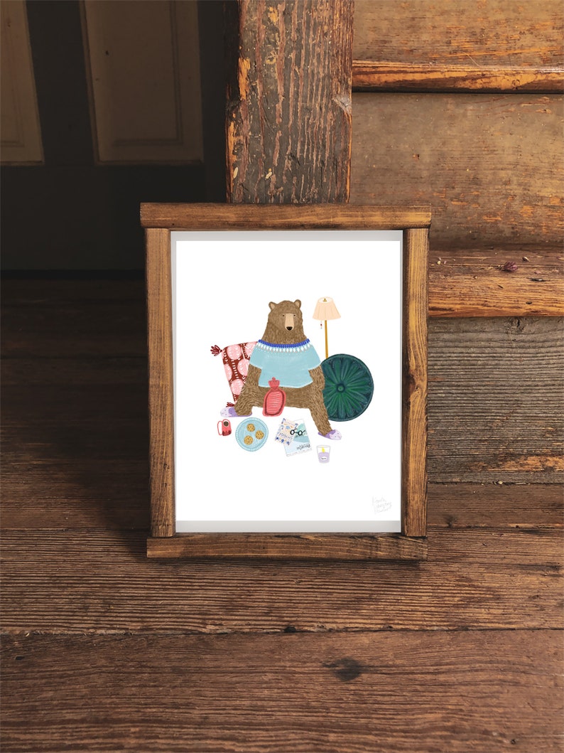 May include: A brown bear wearing a blue sweater and a beaded necklace sits on a patterned pillow with a teal blue round cushion behind it. The bear is holding a red hot water bottle and a glass of water. There are cookies, a red and white striped cup, a blue and white patterned map, and a lamp with a white shade on the white background.
