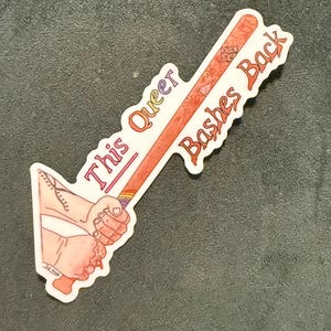Könnte beinhalten: Aufkleber mit einem Baseballschläger-Design und dem Text "This Queer Bashes Back". Der Schläger wird von einer Hand mit einem Regenbogen-Griff gehalten. Der Aufkleber hat einen weißen Rand.