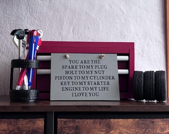 May include: A rectangular gray sign with black text reading: YOU ARE THE SPARK TO MY PLUG, BOLT TO MY NUT, PISTON TO MY CYLINDER, KEY TO MY STARTER, ENGINE TO MY LIFE, I LOVE YOU. It sits before a maroon tool chest with tools and tire-shaped accents.