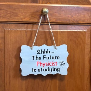 May include: A light gray, scalloped-edge wooden sign hanging from a jute twine cord. The sign features black text that reads "Shhh... The Future Physicist is studying" with the word "Physicist" in red and a small black atom symbol graphic.