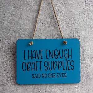 May include: A rectangular bright blue hanging sign with rounded corners and a jute rope hanger. The sign features black text that reads: I HAVE ENOUGH CRAFT SUPPLIES -SAID NO ONE EVER. The sign is suspended from a single clear hook.