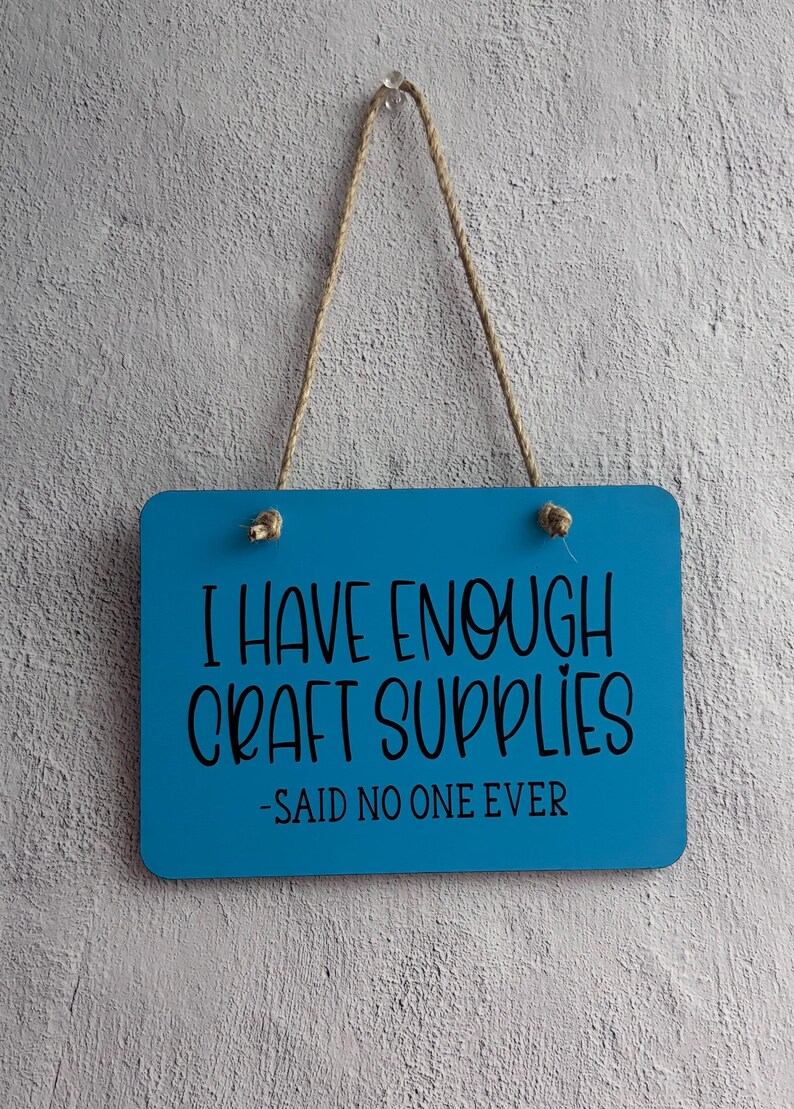 May include: A rectangular bright blue hanging sign with rounded corners and a jute rope hanger. The sign features black text that reads: I HAVE ENOUGH CRAFT SUPPLIES -SAID NO ONE EVER. The sign is suspended from a single clear hook.