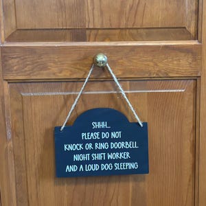 May include: A rectangular black wooden sign with rounded top corners, hanging from a jute twine cord. The sign features white text that reads: SHHH... PLEASE DO NOT KNOCK OR RING DOORBELL. NIGHT SHIFT WORKER AND A LOUD DOG SLEEPING.