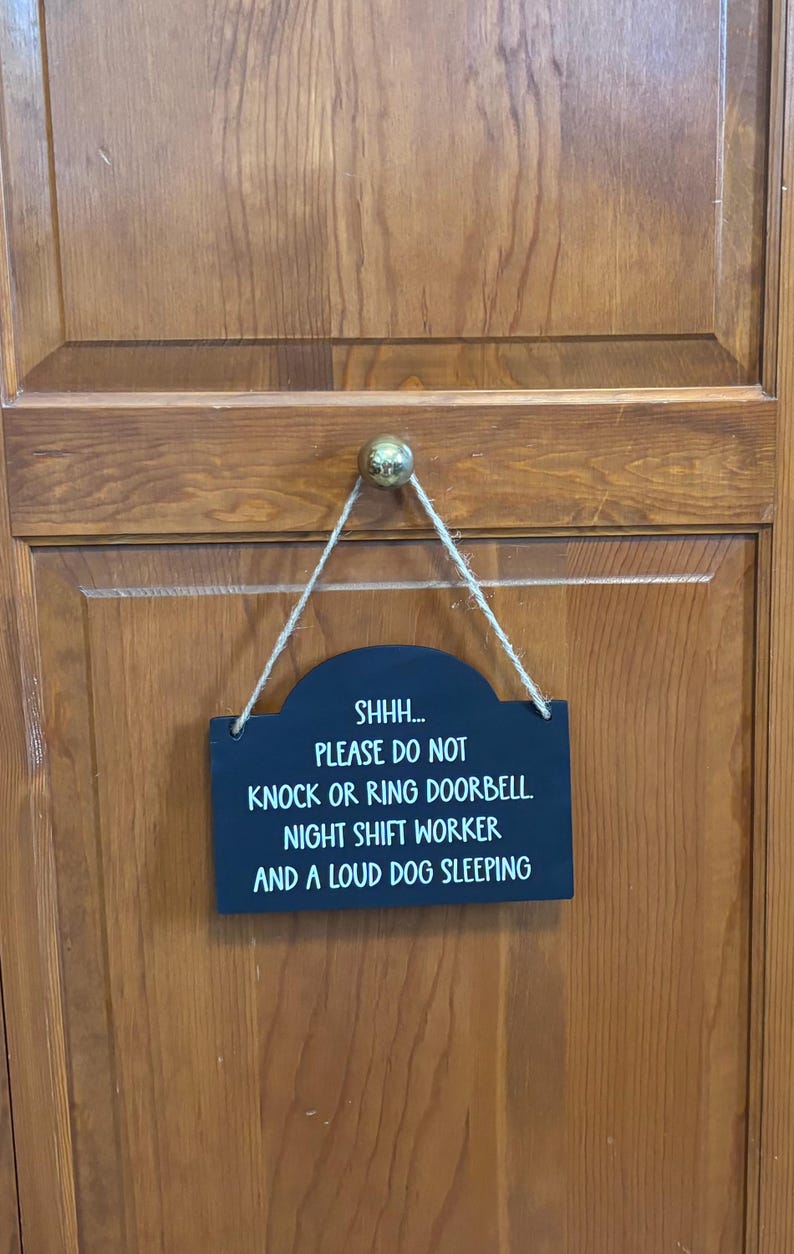 May include: A rectangular black wooden sign with rounded top corners, hanging from a jute twine cord. The sign features white text that reads: SHHH... PLEASE DO NOT KNOCK OR RING DOORBELL. NIGHT SHIFT WORKER AND A LOUD DOG SLEEPING.