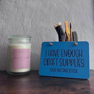 May include: A clear glass candle jar with a pink label reading GOOD VIBES SOOTHE and a blue rectangular sign with the black text I HAVE ENOUGH CRAFT SUPPLIES -SAID NO ONE EVER, holding several paintbrushes in a back compartment.