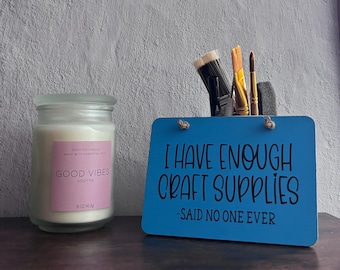 May include: A clear glass candle jar with a pink label reading GOOD VIBES SOOTHE and a blue rectangular sign with the black text I HAVE ENOUGH CRAFT SUPPLIES -SAID NO ONE EVER, holding several paintbrushes in a back compartment.