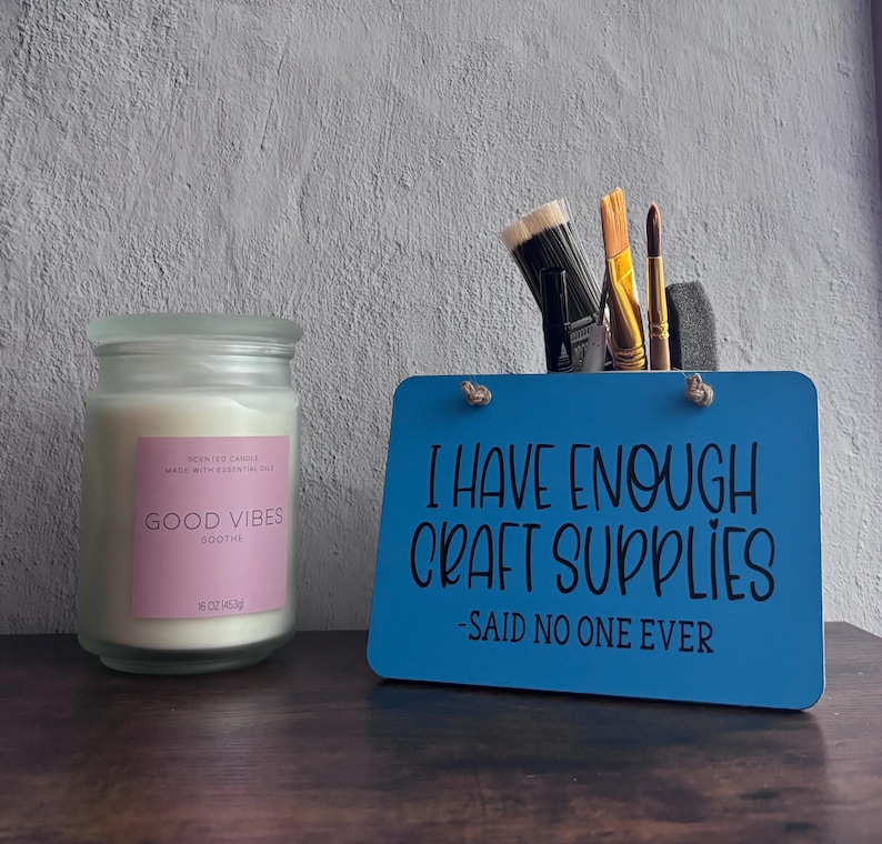 May include: A clear glass candle jar with a pink label reading GOOD VIBES SOOTHE and a blue rectangular sign with the black text I HAVE ENOUGH CRAFT SUPPLIES -SAID NO ONE EVER, holding several paintbrushes in a back compartment.