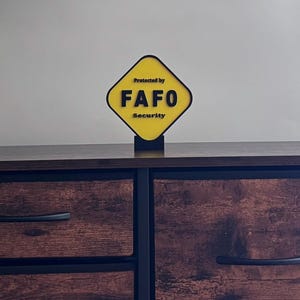 May include: A yellow and black sign that reads "Protected by FAFO Security." The sign is diamond-shaped with rounded corners and is mounted on a black base. The sign is placed on a dark brown wooden cabinet.