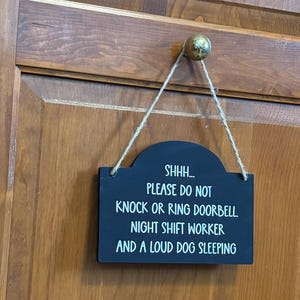 May include: A rectangular black sign with rounded top corners, hanging from a brass knob by a jute twine string. The sign features white text that reads: SHHH... PLEASE DO NOT KNOCK OR RING DOORBELL. NIGHT SHIFT WORKER AND A LOUD DOG SLEEPING.