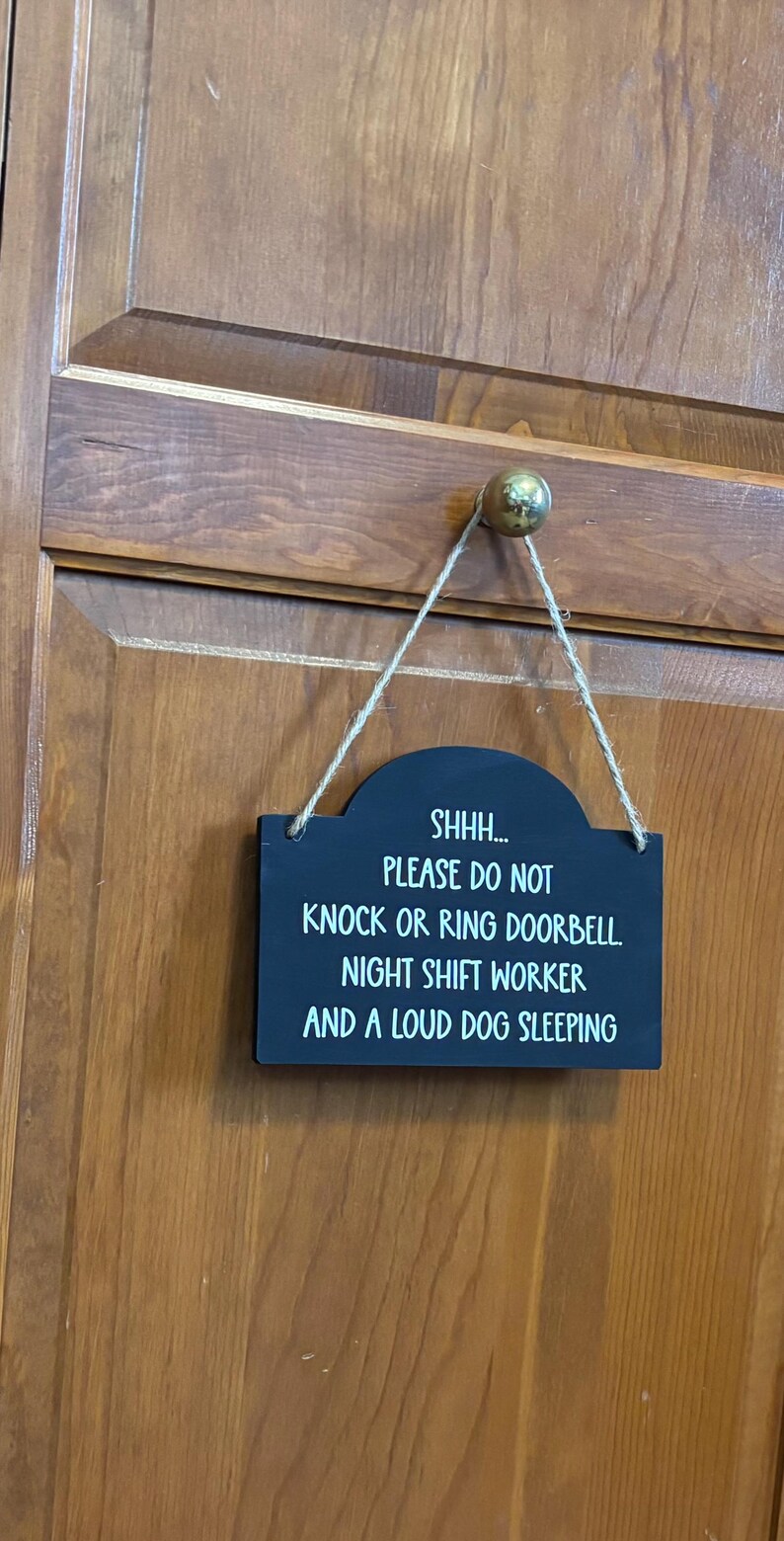 May include: A rectangular black sign with rounded top corners, hanging from a brass knob by a jute twine string. The sign features white text that reads: SHHH... PLEASE DO NOT KNOCK OR RING DOORBELL. NIGHT SHIFT WORKER AND A LOUD DOG SLEEPING.