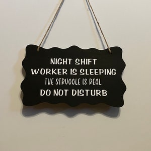 May include: Black chalkboard sign with white text that reads "Night Shift Worker is Sleeping, The Struggle is Real, Do Not Disturb". The sign is hanging from a string.
