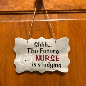 May include: A white wooden sign with a scalloped edge hangs from a string. The sign reads "Shhh... The Future NURSE is studying". A white nurse's cap with a red cross is pictured next to the word "NURSE".
