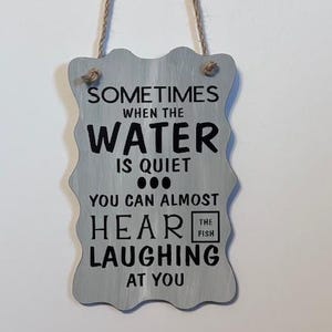 May include: A gray, wavy-edged sign with black text that reads "Sometimes when the water is quiet... you can almost hear the fish laughing at you." The sign is suspended by a natural twine cord.
